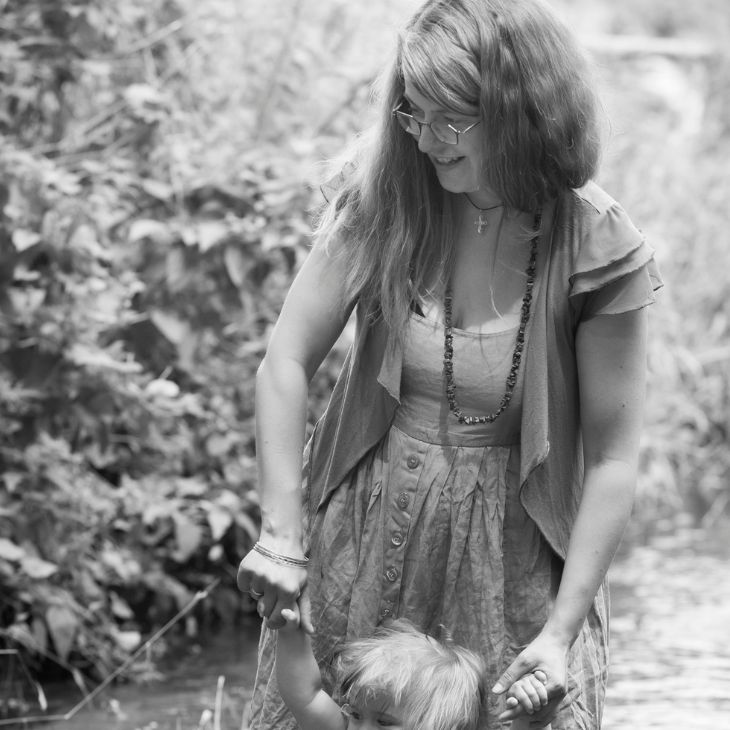 ein Schwarz-weiß-Bild im Hintergrund Büsche eine Frau mit langen Haaren, Brille und einem Kleid hält lächeln ein kleines Kind an den Händen von dem Kind ist nur der Haarschopf zu sehen