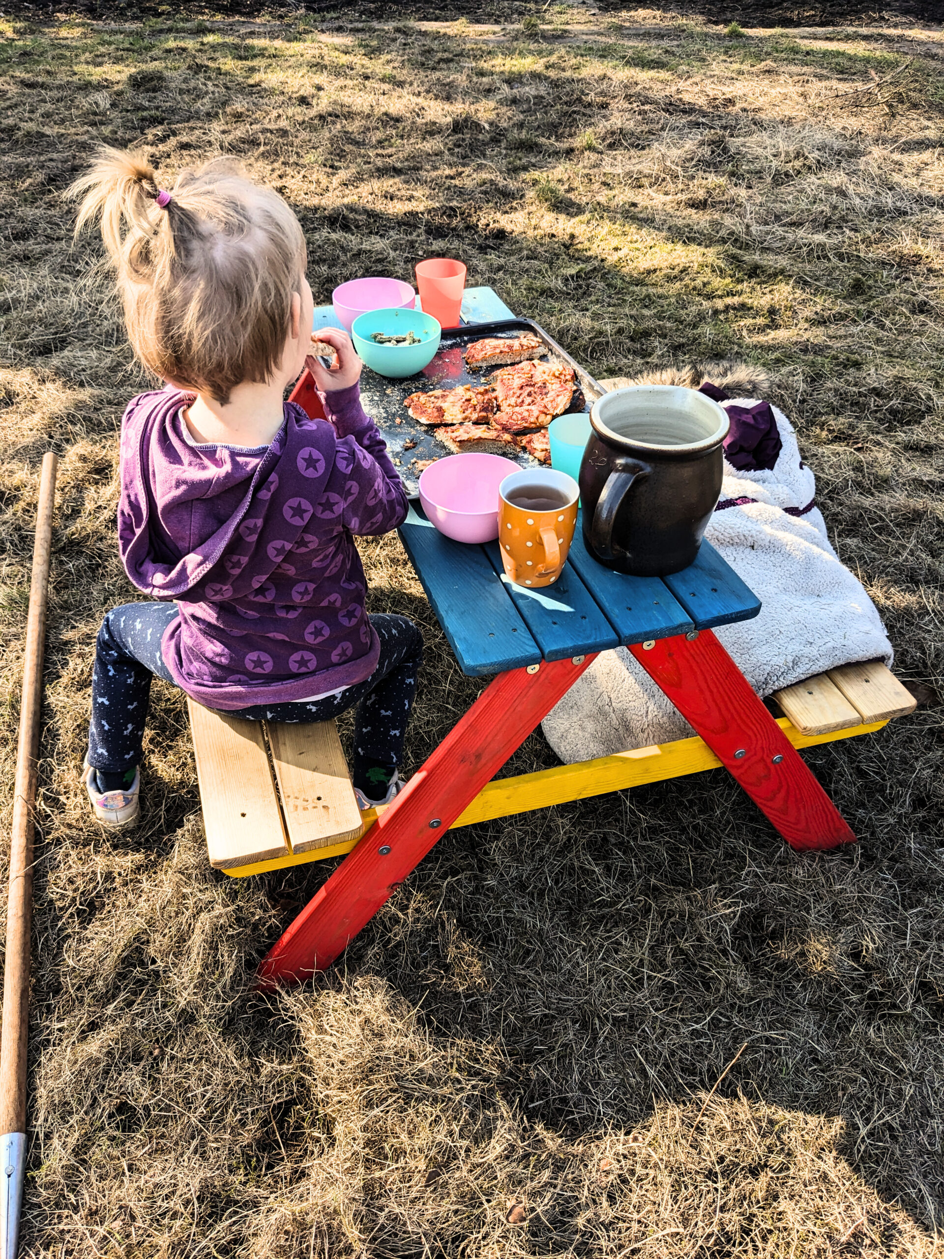 Ein Kind sitzt an einem kleinen, bunten Picknicktisch, auf dem ein Krug und mehrere bunte Becher und Schüsseln stehen