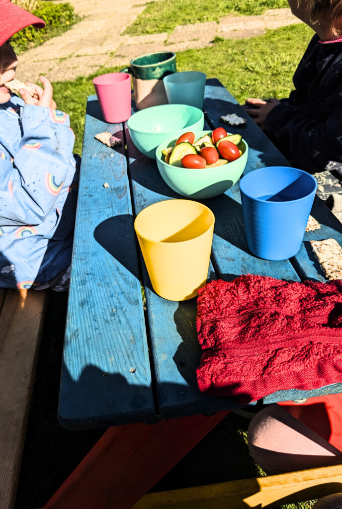Ein bunter Kindertisch auf dem einige Becher und Schüsseln stehen. Kinder sitzen an dem Tisch und essen